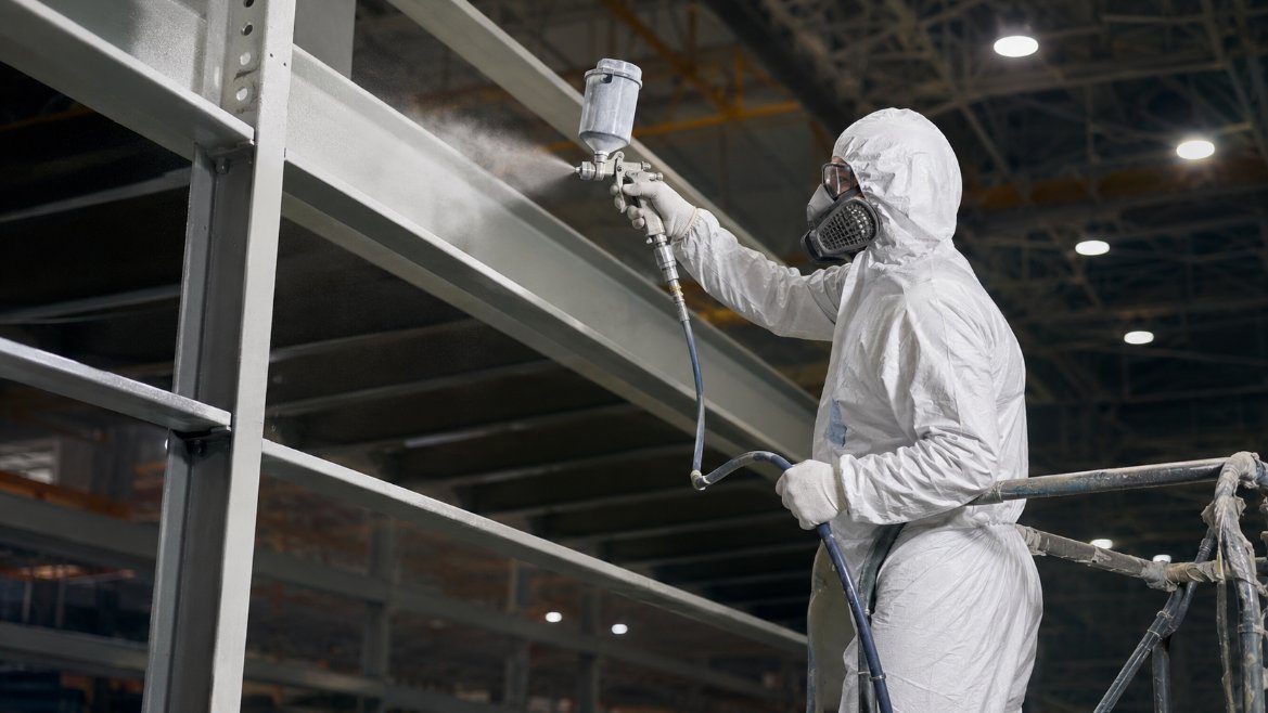 Pintor industrial con EPI completo aplicando pintura a pistola airless en interior de nave